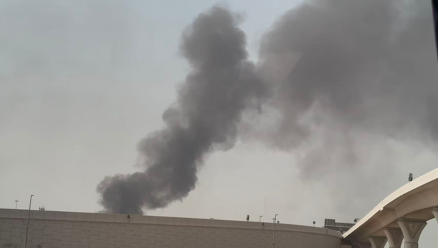 Thick black smoke rises above a large, flat-roofed building under a hazy sky, with part of a white elevated structure visible on the right side.